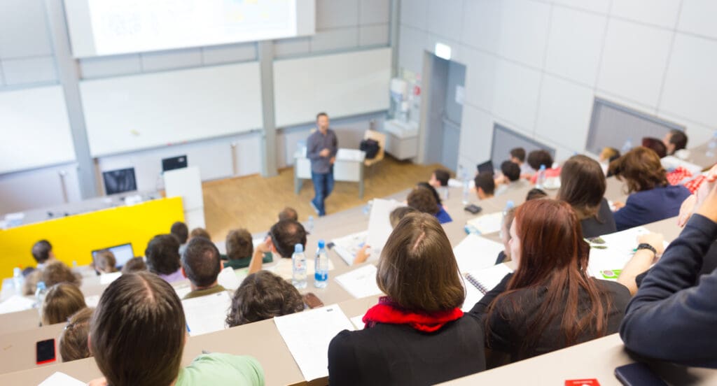 Speaker giving presentation in lecture hall at university. Students listening to lecture and making notes.