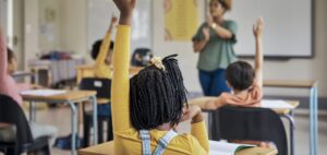 Kid in class, image from behind. They are sitting at a desk with their hand raised. They have a yellow shirt and dark brown hair.