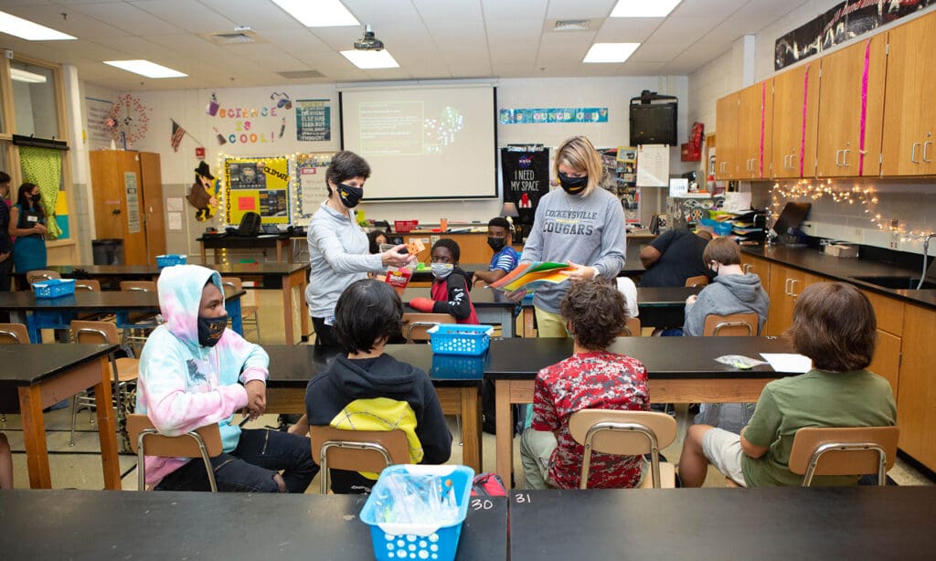 class room with students seating and teachers standing, in a circle with desks around them and everyone is wearing a mask