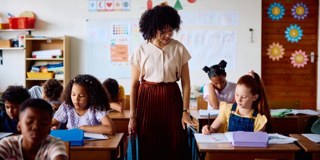 Female teacher in her 20s standing beside desk of schoolgirl, looking at her work in class